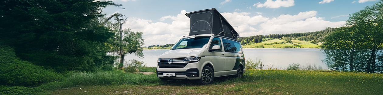 Ein VW California steht mit ausgeklappten Dach an einem See